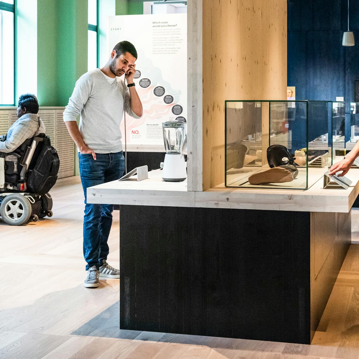 Photograph of an exhibition gallery space. In the background to the right is a blue stained wood wall and to the left a light green painted wall containing windows. To the front of the frame is a display case table, divided close to the middle by a vertical wooden wall. On the right side a woman is looking at objects in a glass case whilst touching an info panel with her right hand. On the left side a man is holding a single headphone to his left ear, listening to an audio commentary. In front o