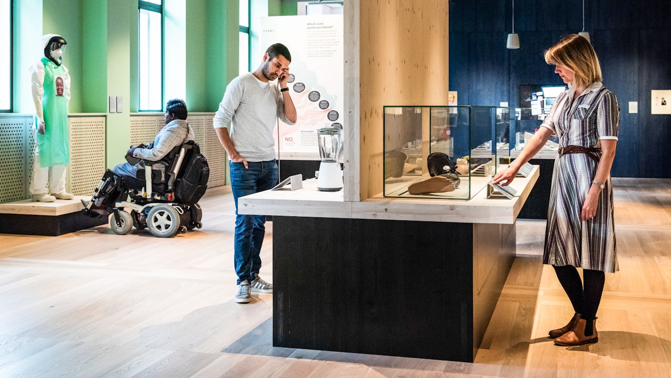 Photograph of an exhibition gallery space. In the background to the right is a blue stained wood wall and to the left a light green painted wall containing windows. To the front of the frame is a display case table, divided close to the middle by a vertical wooden wall. On the right side a woman is looking at objects in a glass case whilst touching an info panel with her right hand. On the left side a man is holding a single headphone to his left ear, listening to an audio commentary. In front o