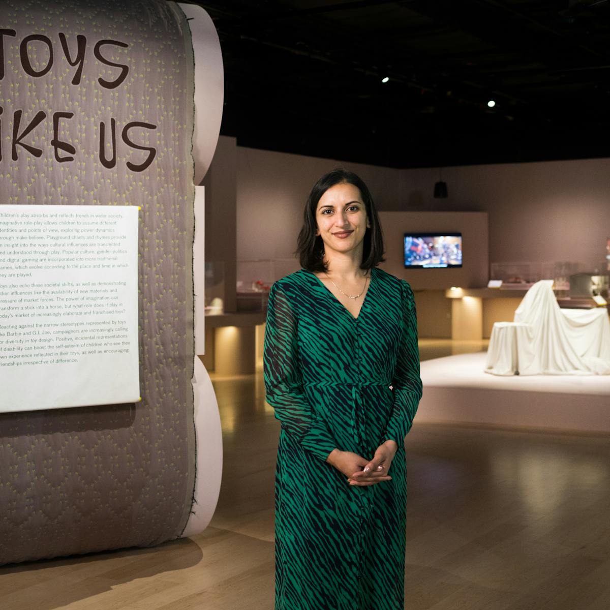 Photograph of a woman standing an exhibition gallery smiling to camera. To the left of her is an exhibition text panel with the title 'Toys Like Us' written above it. In the distance behind her to the right are exhibition exhibits and a tv screen.