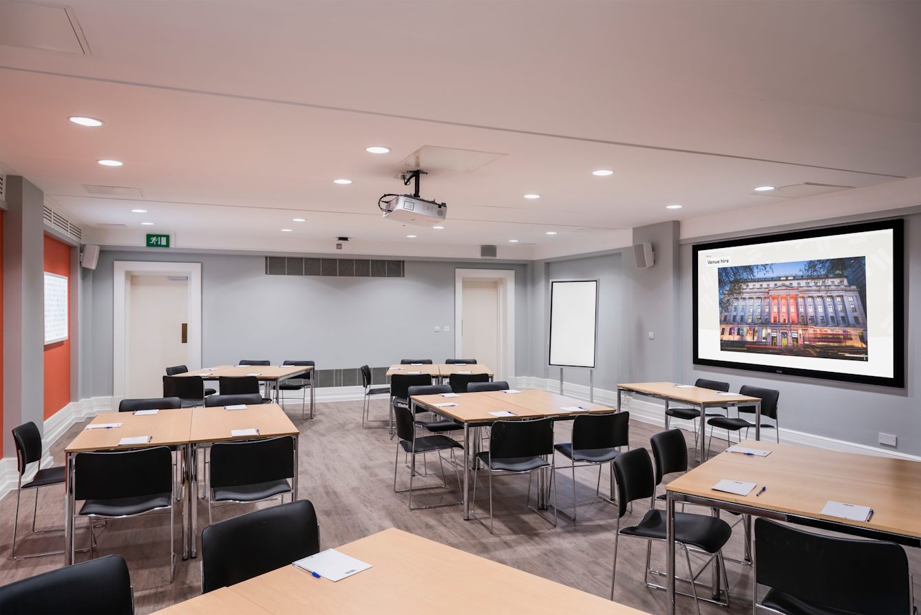 Photograph of the Burroughs room at the Wellcome Collection.
Photograph shows a cabaret set-up, with desks placed in small groups, and a presenting screen and whiteboard at the front of the room.