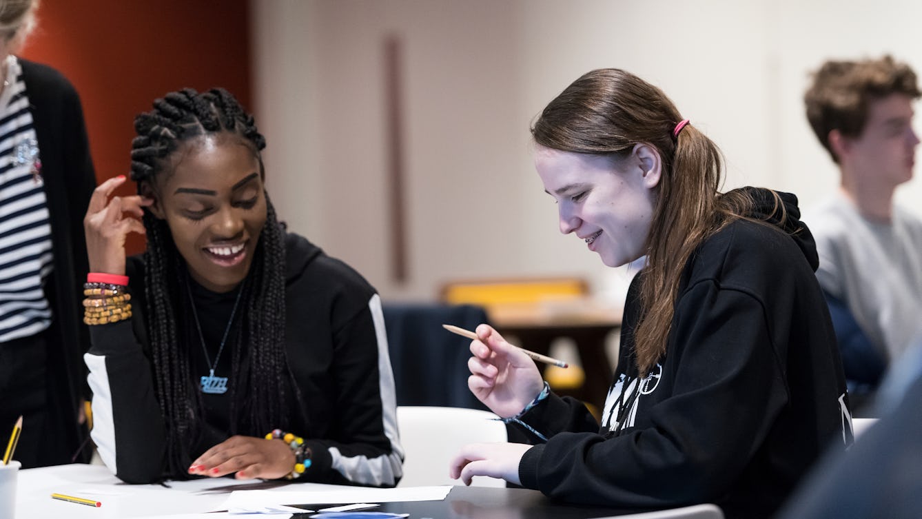 Photograph of two students sat at a table looking down at the activities that they're working on. They are holding a pencil and working on white paper. 