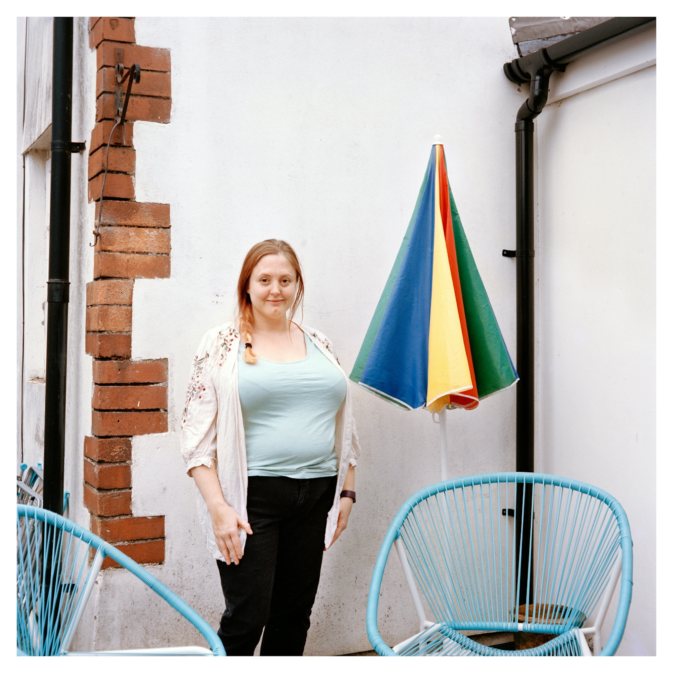 Portrait photograph of Jayne, a woman with blonde and red hair, standing in front of a white painted brick wall. Her hair is in a side braid and she is wearing a light blue t-shirt, a white floral cardigan and black trousers.
Her hands are by her side and she is looking directly at the camera with a slight smile. There is a colourful patio umbrella next to her and several blue beach chairs.