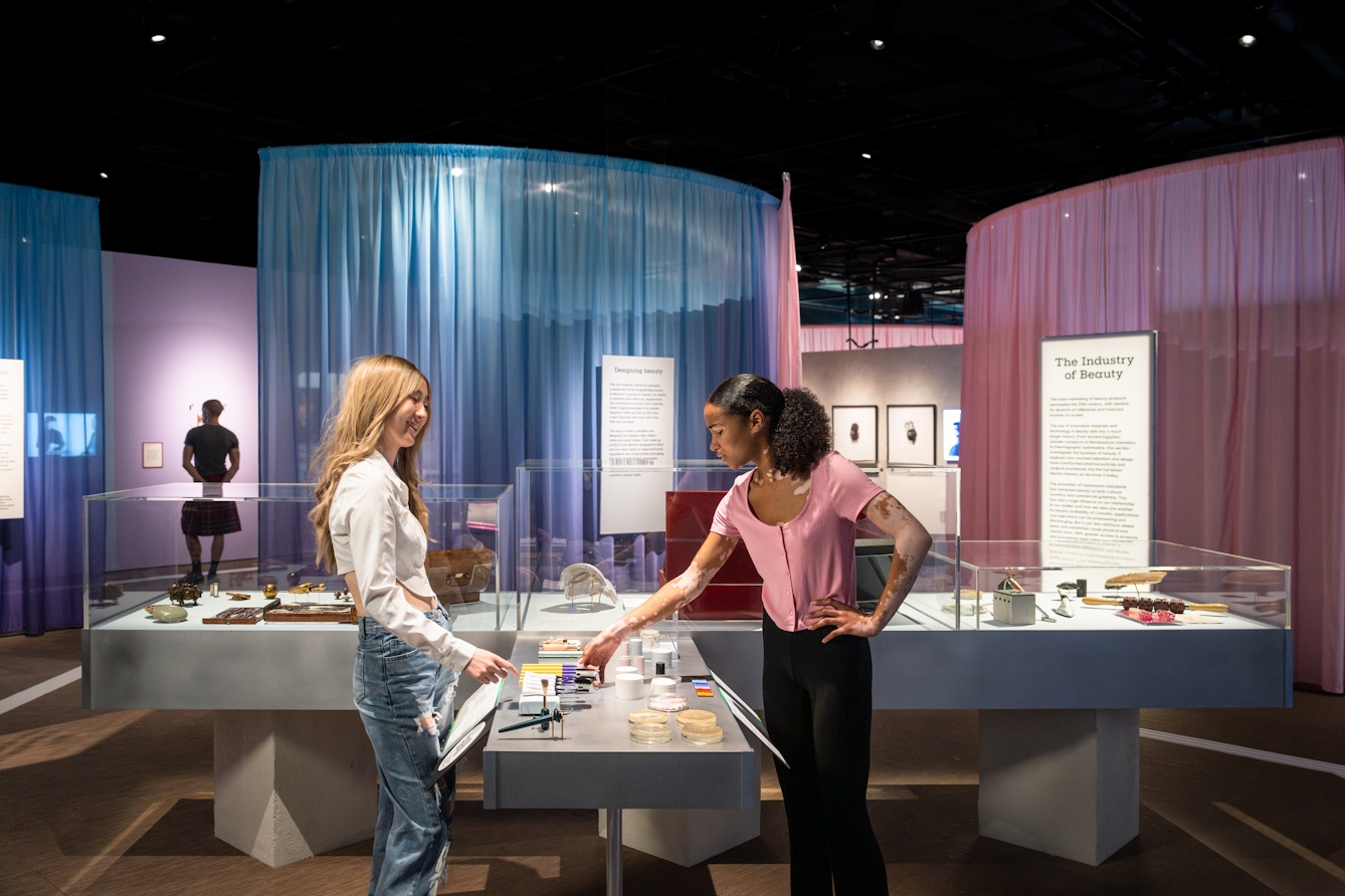 Photograph of 2 gallery visitors stood at an open display case discussing and touching the tactile objects.