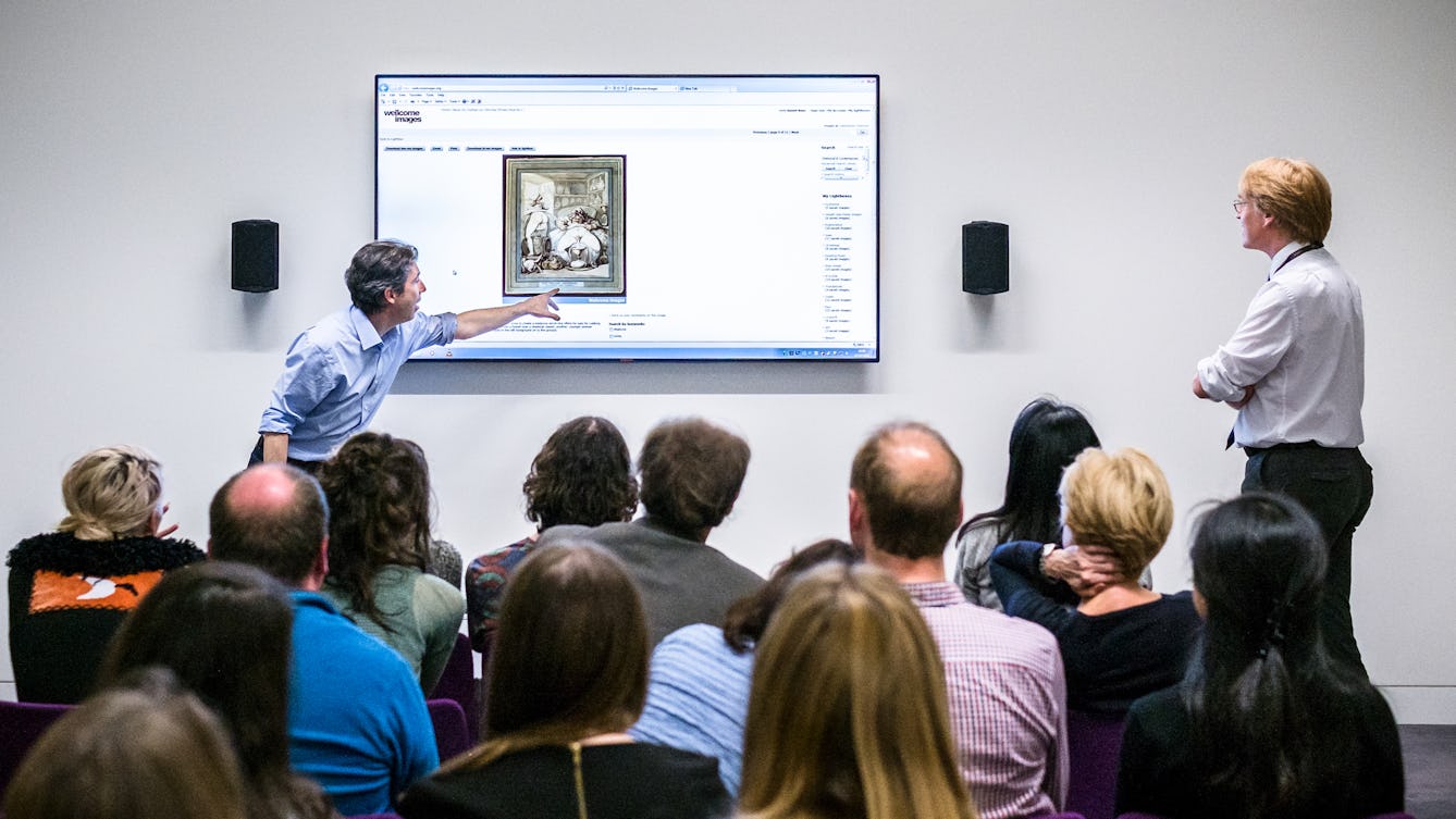 Photograph of a man giving a talk to an audience. He is pointing to a large screen besides him.
