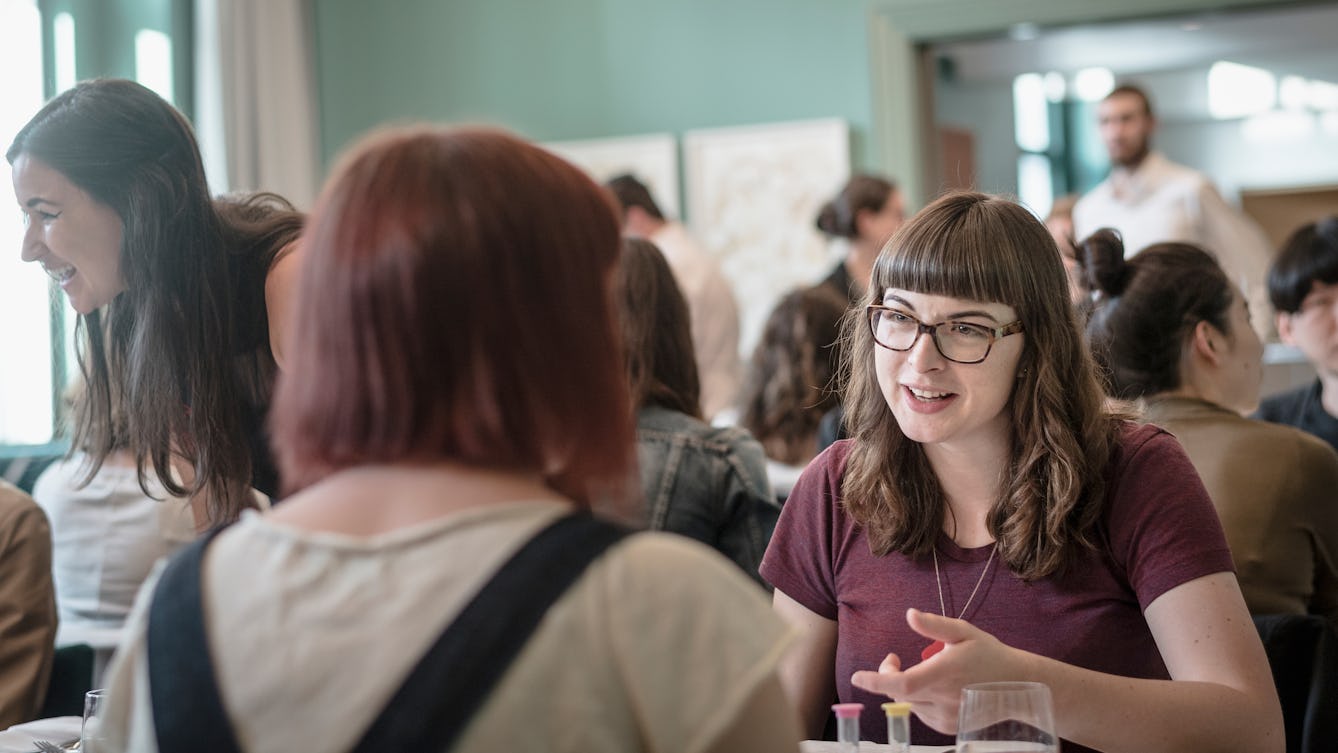 Two people having a conversation across a table in Wellcome Kitchen. Behind them, out of focus is a busy room with people seated at tables.