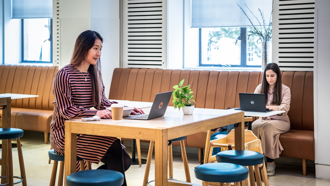 Photograph of a café interior. A woman is sat at a high table working on an open laptop. To her right is a disposable coffee cup and to her left is a small pot plant. In the background another woman is sat on a long bench seat at a table. She is also working on an open laptop