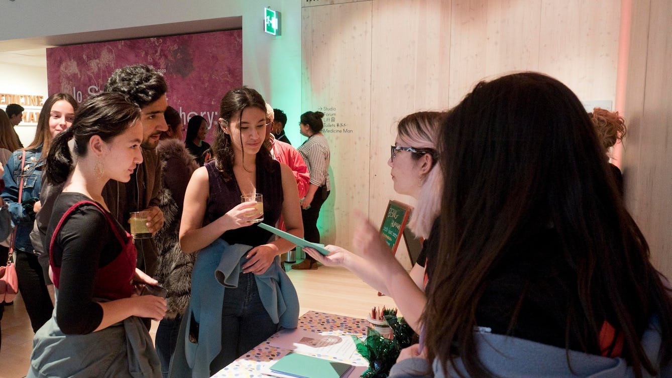 Young people standing outside the Being Human Exhibition at Wellcome Collection. That are holding mocktails and interacting with staff at a table who are handing  visitors paper exhibition guides.