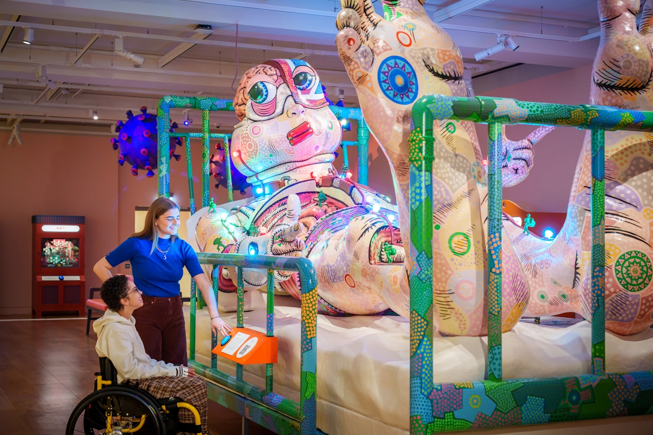 Photograph of a large sculpture in an exhibition space. The sculpture is a large person covered in colourful painted patterns, lying on a hospital bed. Two gallery visitors to the side of the bed are activating the button which sets off a light show covering the surface of the sculpture.