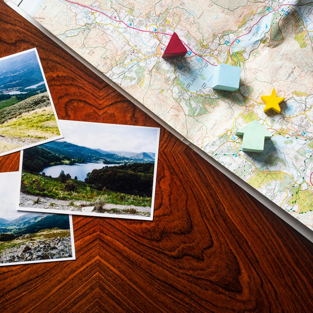 Photograph of a warm wood grained, polished tabletop from above. Resting on the table, cutting diagonally across the top right corner is part of an unfolded map showing roads, lakes, and the contours of hills. Resting on the map are four three dimensional shapes, a red triangle, a light blue cube, a bright yellow star and a light green arrow. To the centre and bottom left of the image are three photographic prints with thin white borders. These photographs show mountain walking scenes looking over hills, rocky outcrops, valleys and lakes. In one of the photographs a lone figure can be seen looking out at the view.