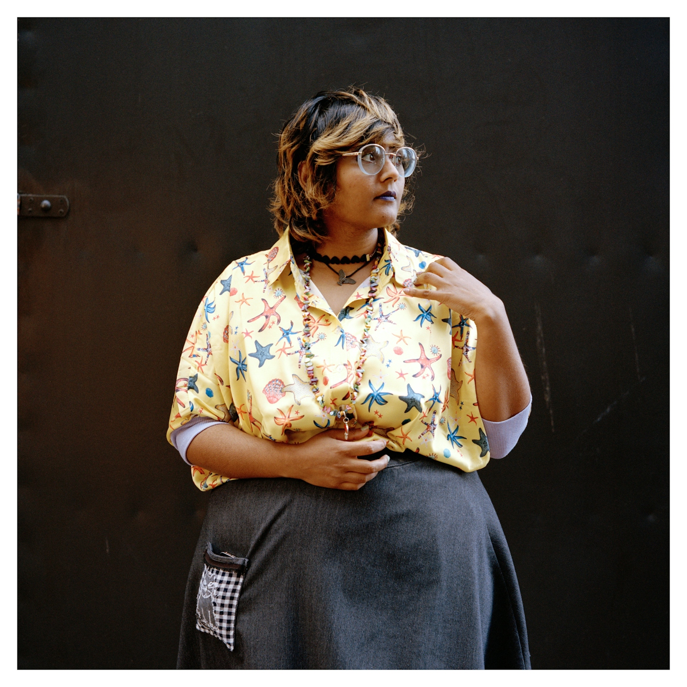 Portrait photograph of Sumita, a woman with shoulder length brown hair with blonde streaks. She is wearing a yellow shirt with a starfish pattern and a grey skirt. She is wearing glasses, purple lipstick and several chunky necklaces.
She is standing in front of a brown backdrop and is looking to her left, with a neutral expression.