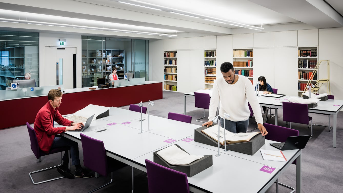 Photograph of a library rare materials reading room. It is a wide shot which shows two large tables, an enquiry desk and shelving along the wall containing books. At the table in the foreground a man is sitting working on a laptop computer. Another man is standing leaning over a book support which has a fragile book resting on top of it. In the background is another reader sat at the far table.