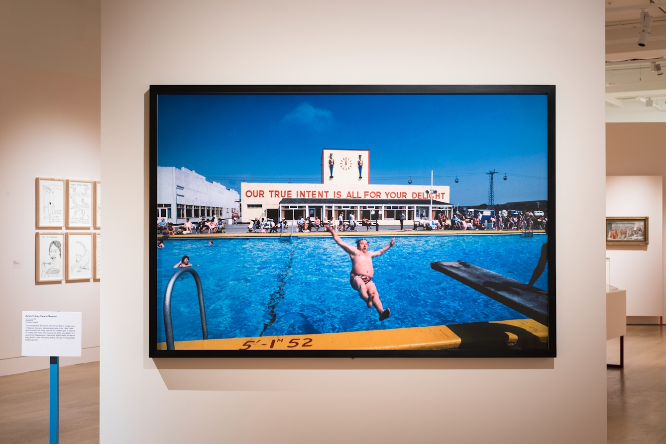 Photograph of an exhibition gallery space showing a section of light pink coloured wall, on which is hung a large colour photographic print. The print shows a sunny holiday camp scene with a large swimming pool in the foreground and a 1930 building in the background with the large words 'Our true intent is all for your delight' across the front. In the foreground a man wearing swimming trunks has been photography in mid air as he falls backwards into the pool. Behind the section of wall the rest of the gallery space can be seen in the distance.
