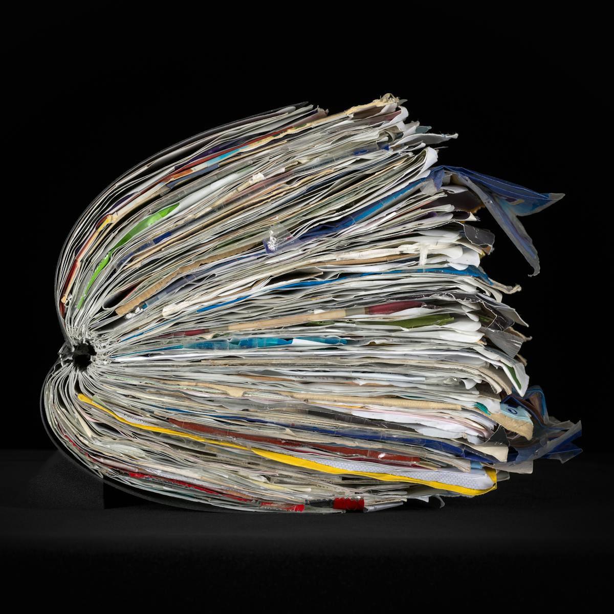 Side view of a large ring-bound scrapbook on a black background. The scrapbook is bulging with ephemera pasted within it.