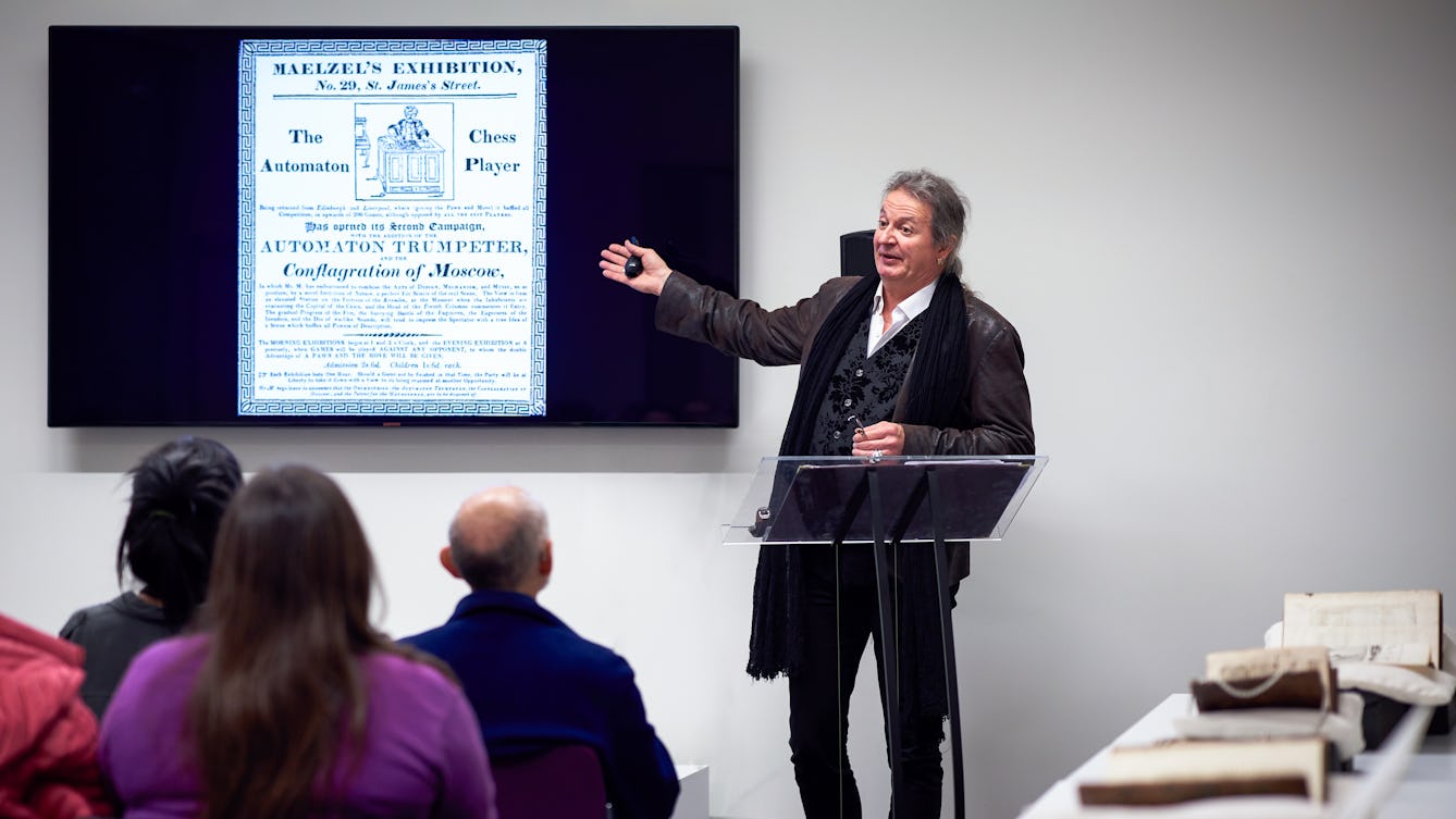 Photograph of a made standing at a lectern beside a large screen giving a talk to a seated audience.