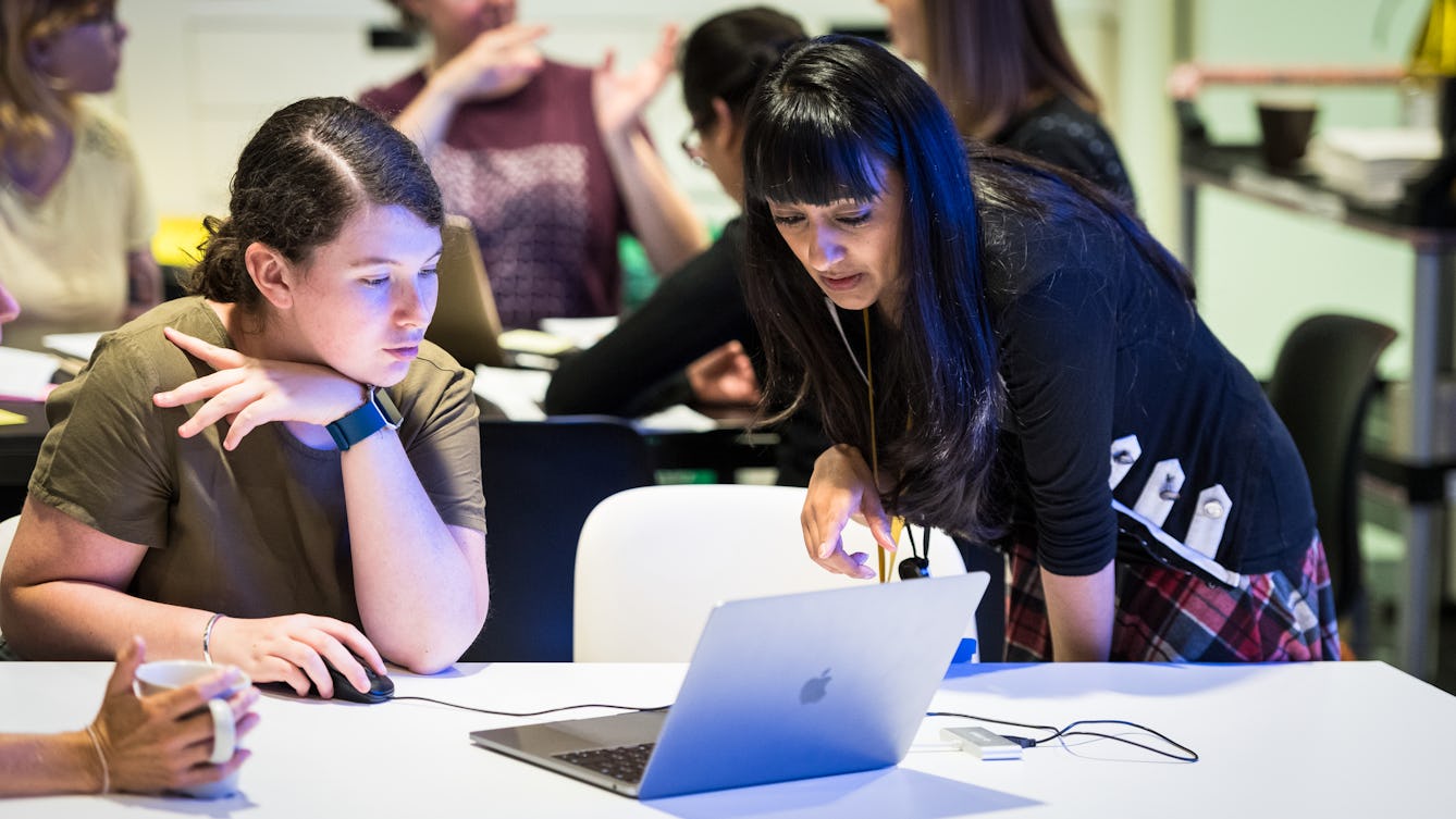 Photograph of two young women in a classroom environment. One is sat at a table holding a computer mouse and looking at a laptop screen. The other is standing, leaning over the table looking and point at the same laptop screen.