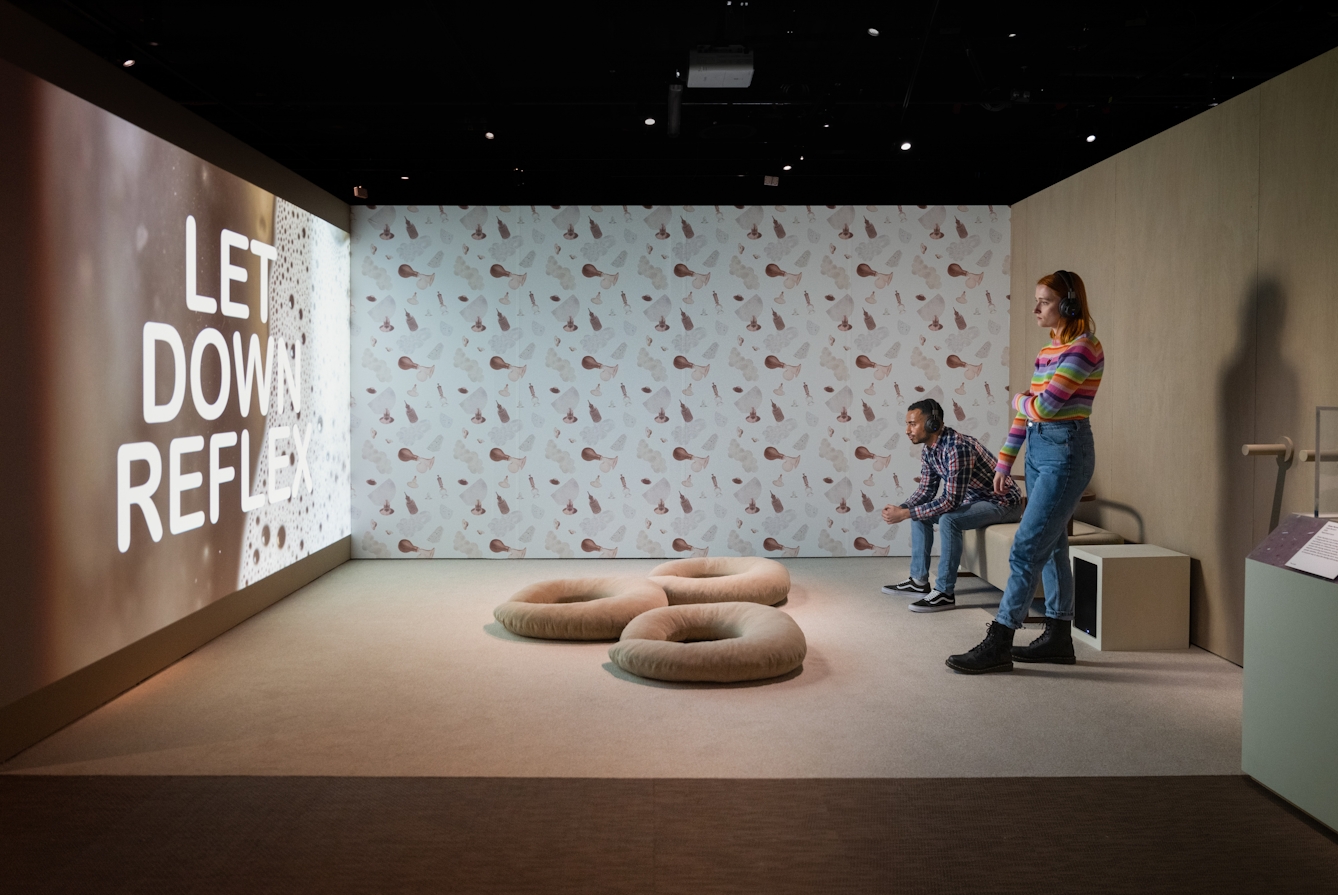 Photograph of 2 exhibition visitors looking at a large projected video in an exhibition gallery setting. One visitor is sat on a bench whilst the other stands to one side. The wall behind them is covered I patterned wallpaper and on the floor are 3 hooped cushion which resemble nursing pillows. On the screen is projected the words, 'Let down reflex'.