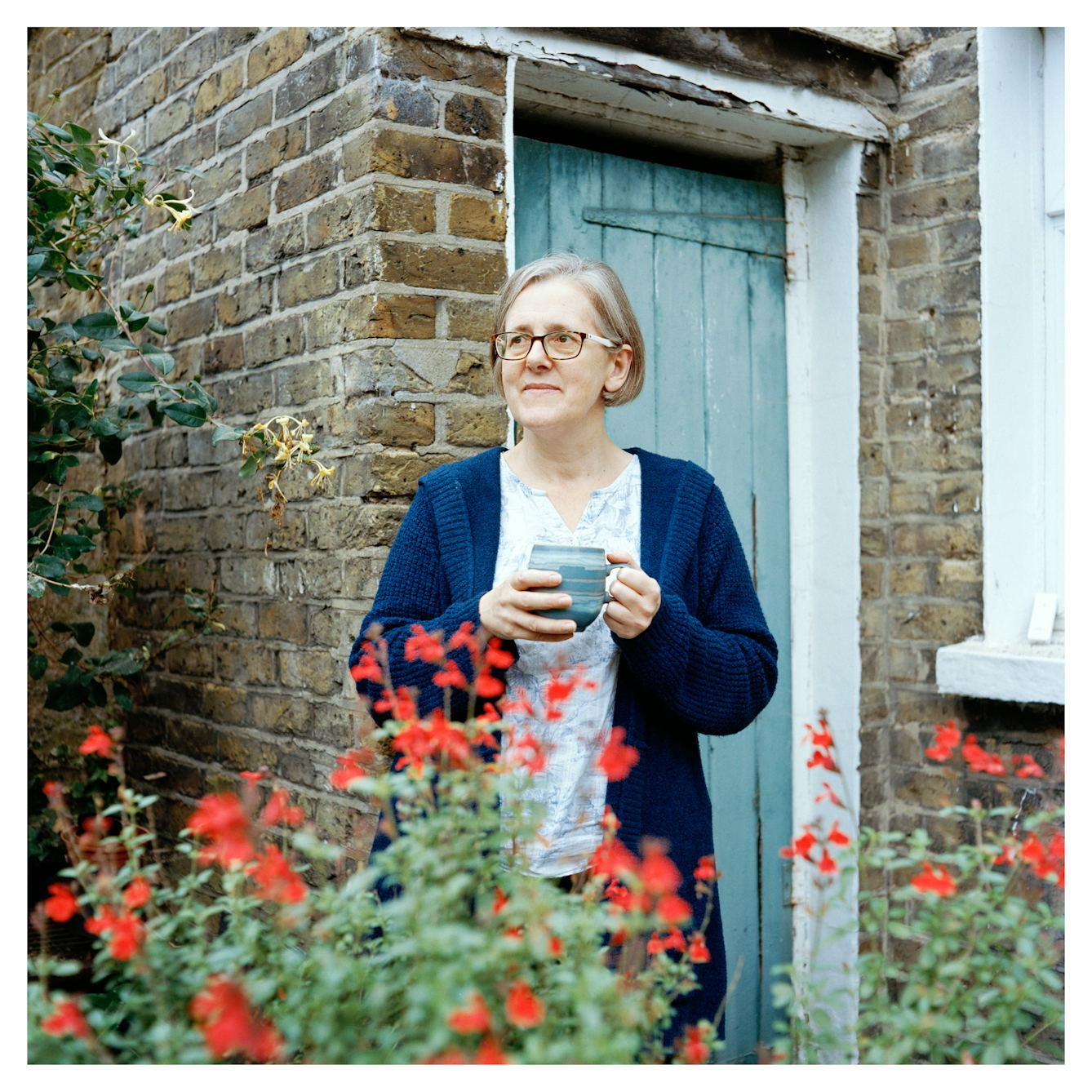 Portrait photograph of Emily, a woman with short grey hair wearing glasses. She is wearing a patterned top and a long blue cardigan and is holding a mug with both hands.
She is standing in front of a brick building with a blue painted wooden door. She is looking into the distance and in front of her are some red wildflowers.