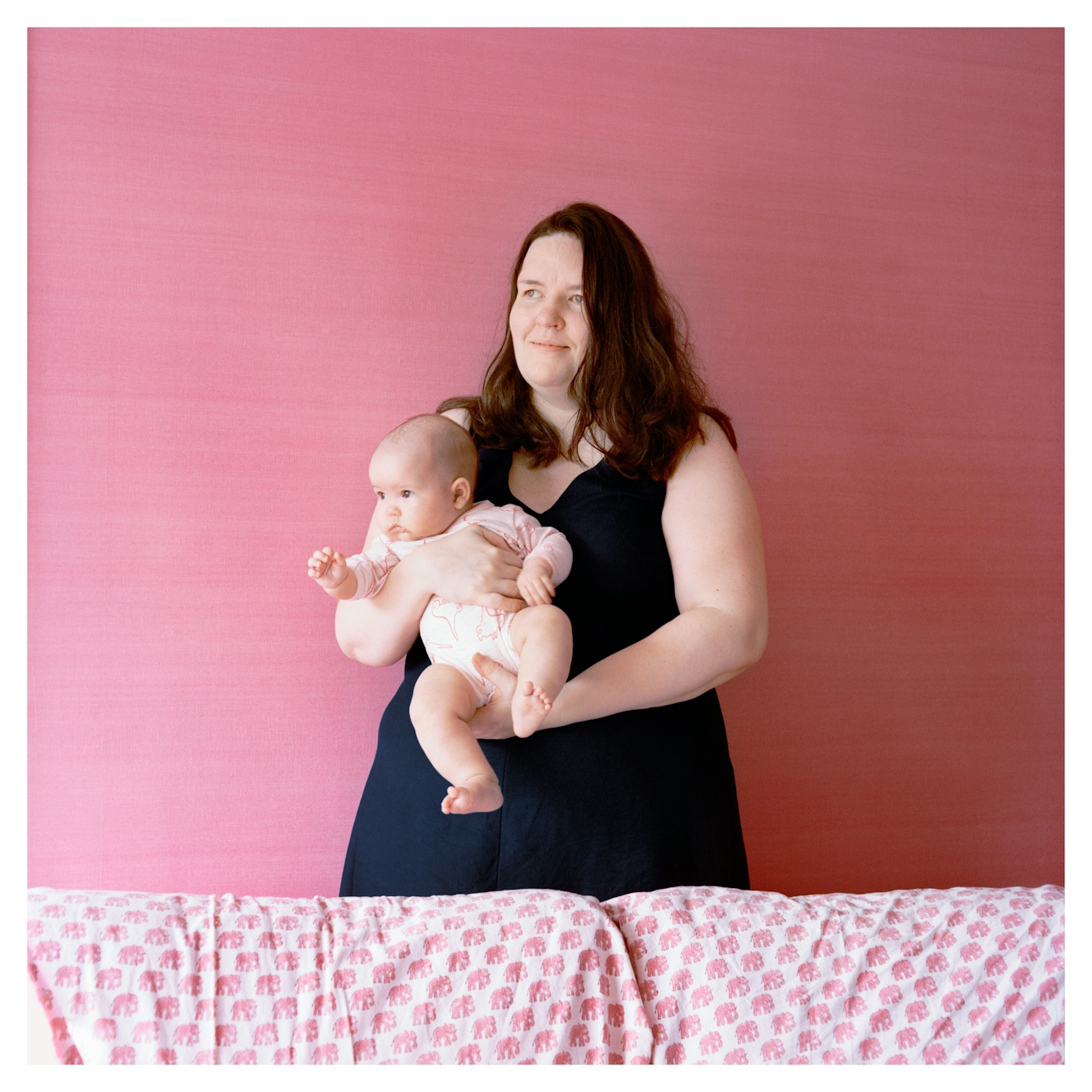 Portrait photograph of Elinor, a woman with shoulder length brown hair, standing in front of a painted pink wall. She is wearing a navy dress and holding a small baby, who is wearing a pink babygro. They are both looking to to the right and Elinor is smiling slightly. There is a patterned white and pink sofa in front of them.