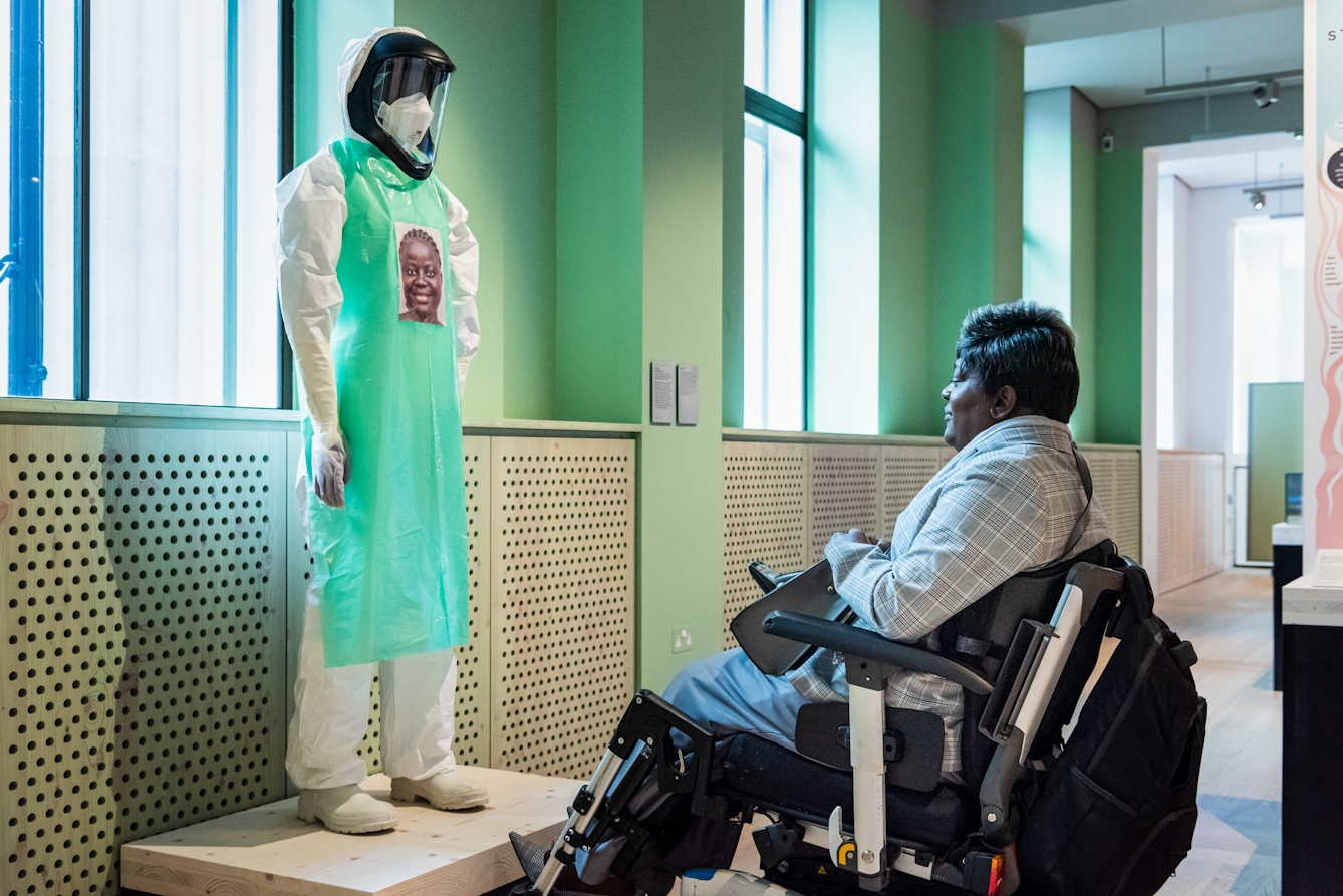 A photograph showing a visitor seated in a wheelchair looking at a model wearing a white hazmat suit, visor and green apron standing on a low wooden platform. On the green apron there is a stickered portrait of the carer that would have been wearing the suit whilst treating Ebola patients.