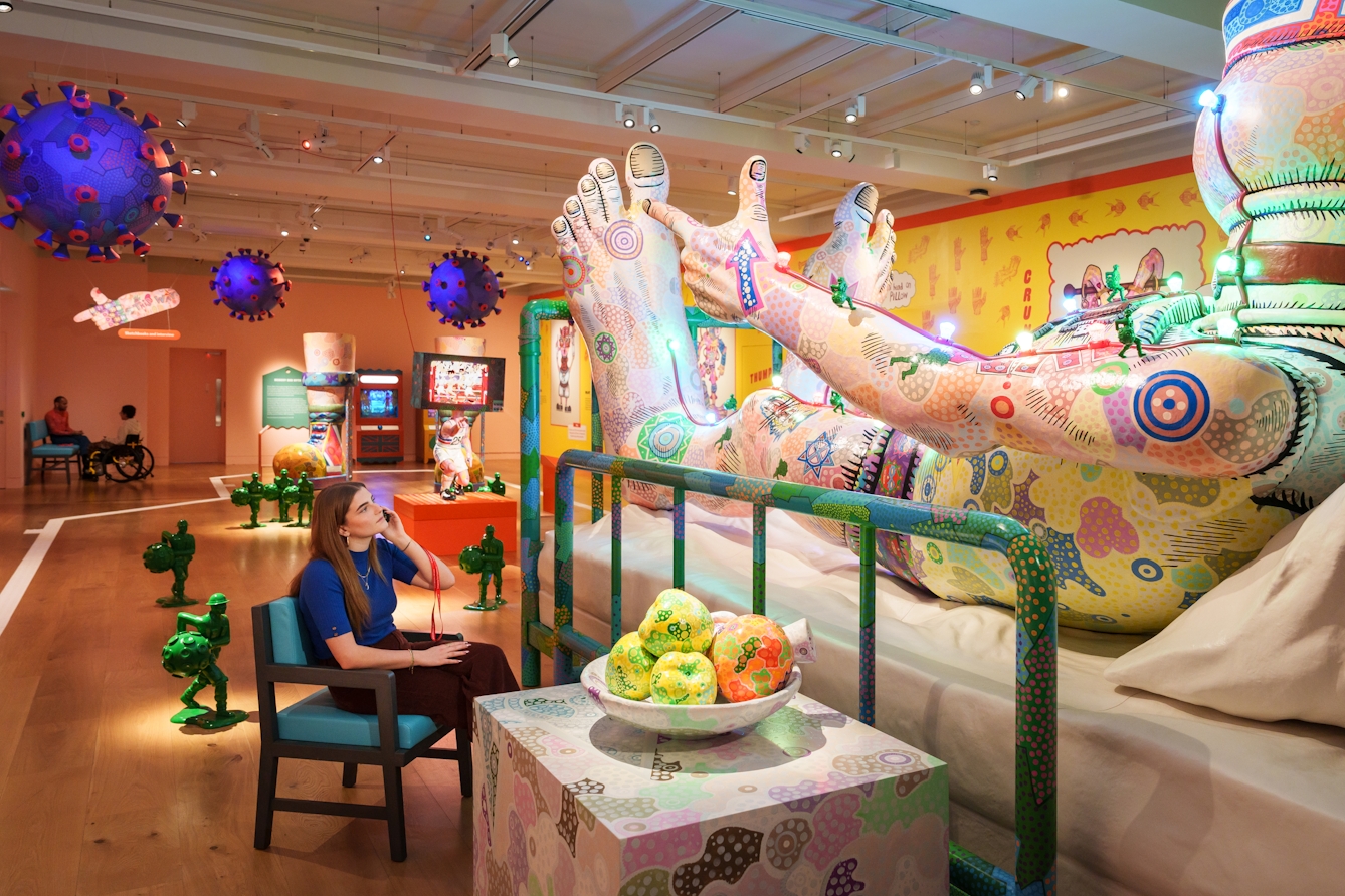 Photograph of a large sculpture in an exhibition space. The sculpture is a large person covered in colourful painted patterns, lying on a hospital bed. A gallery visitor sits beside the bed listening to an audio guide.

