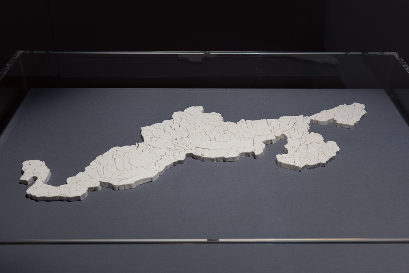 Colour photograph of a white porcelain sculpture of part of a glacier. The porcelain has many cracks in it. The sculpture is in a display cabinet with a glass cover over it. 