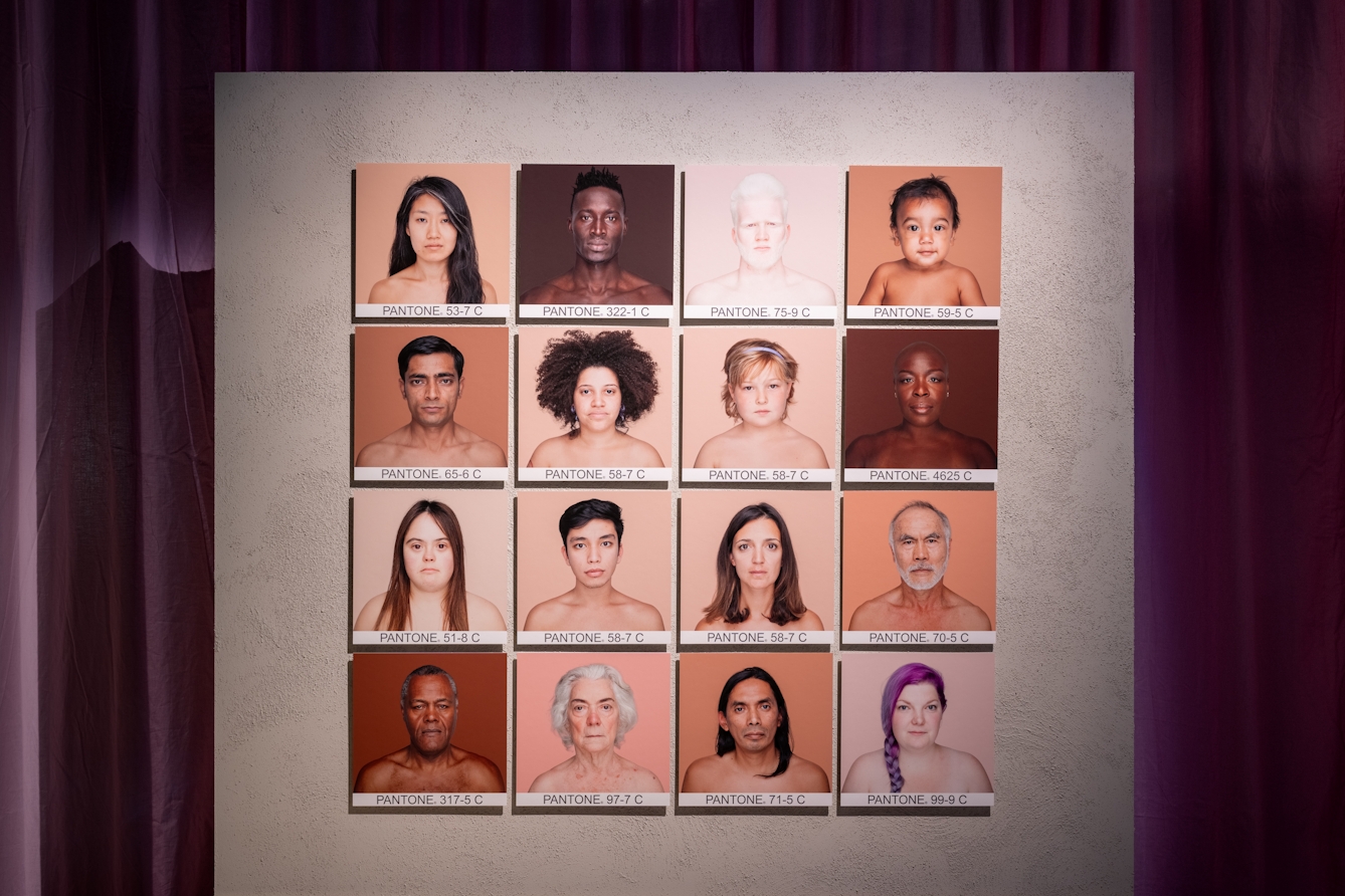 Photograph of a grid of 4 by 4 square portraits of 16 people's head and shoulders. The background colour for each portrait matches the hues of the person's skin. Under each image is a Pantone reference.