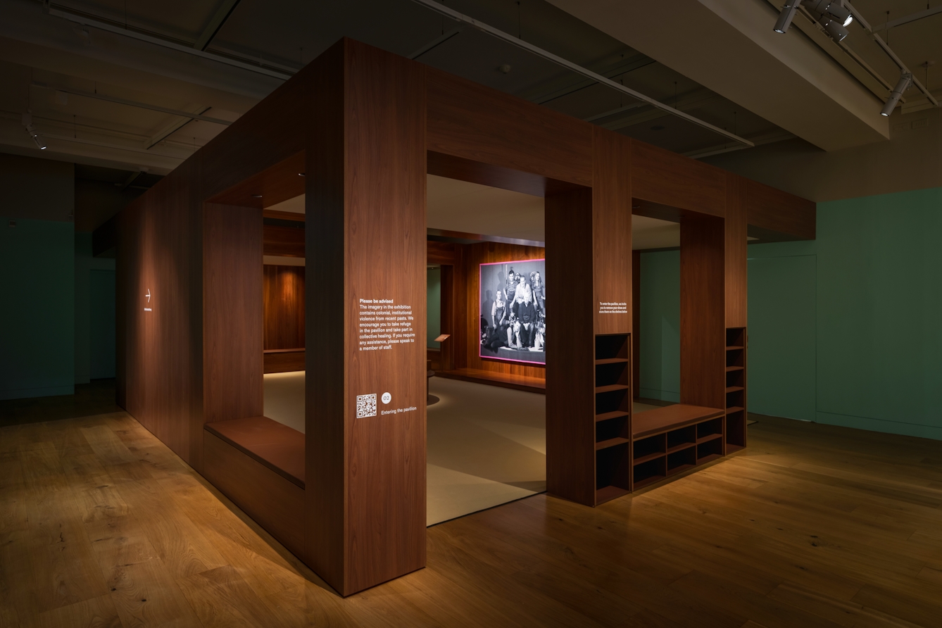 Photograph of a gallery exhibition space showing large tapestry hung on a wooden wall. The tapestry is made up of a black and white, archive photograph, from the late 20th century showing a group of white men and women gathered in a group within an exhibition exhibit. The tapestry is framed with a bright pink border. In surrounding the tapestry is a wooden pavilion-like structure made up of columns, alcoves and seating areas. The walls are olive green in hue and the overall sense is one of a clam reflective space.
