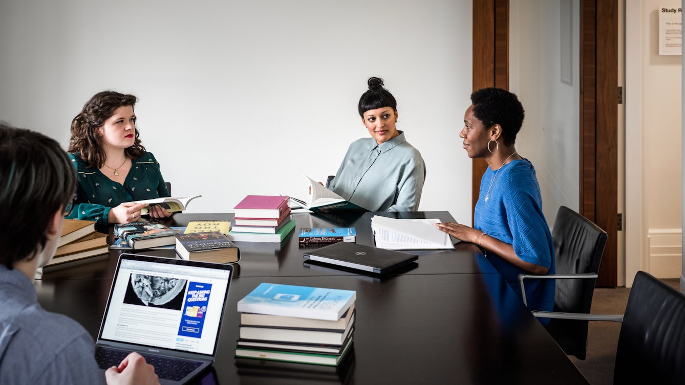 Photograph of four people sat around a large black table in a plain white walled room. In teh background the open wooden door can be seen. On the table are piles of books, a closed laptop computer and an open laptop computer with an website loaded on the screen. The group of people are in a discussion.