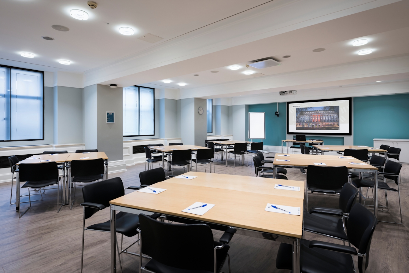 Photograph of the Franks room at the Wellcome Collection.
Photograph shows a cabaret set-up with desks placed together in small groups. There is a projector and screen at the front of the room and several large windows.