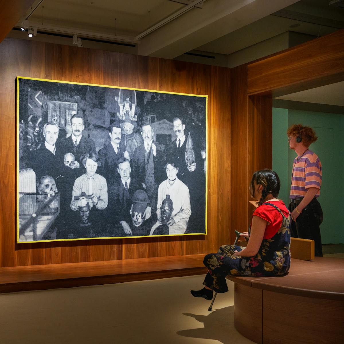 Photograph of a gallery exhibition space showing large tapestry hung on a wooden wall. The tapestry is made up of a black and white, archive photograph, from the early 20th century showing a group of white men and women gathered in a group holding museum artefacts in their hands or on their laps. They are surrounded by other museum exhibits and display cases. The tapestry is framed with a bright yellow border. Surrounding the tapestry is a wider wooden structure containing seating and alcoves. To the right of the image a gallery visitor reclines in one of the alcoves with her back to the tapestry, in contemplation. To the right two more visitors, one sitting, one standing are looking at the tapestry. The standing visitor is listening to an audio guide on headphones. The hues of the scene are warm browns and olive greens.
