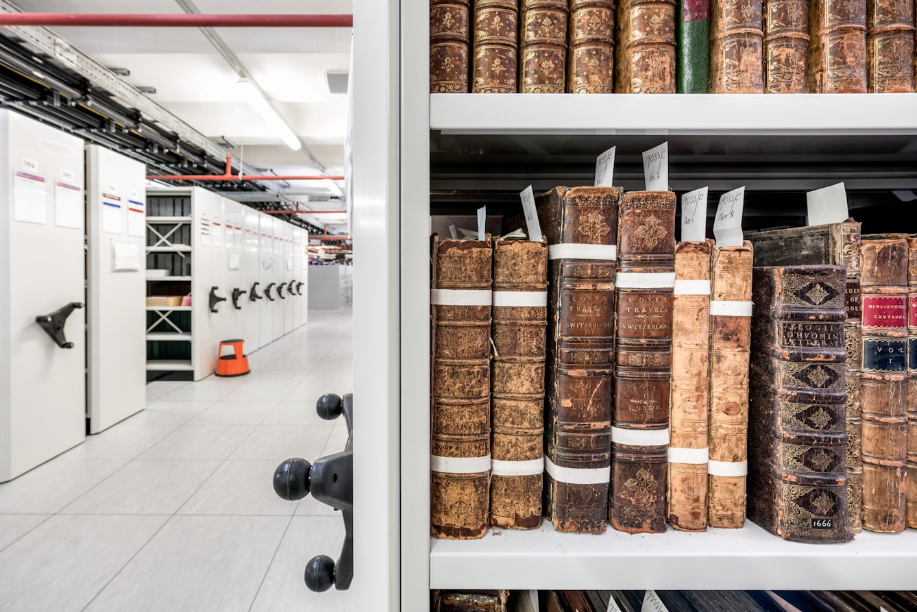 Photograph of a windowless library store containing rolling shelving. The right hand side of the image contains a close up view of a book shelf showing the spines of several fragile books. The left of the image show the corridor between the shelves receding into the distance, an orange kick stool and the utility piping suspended from the ceiling.