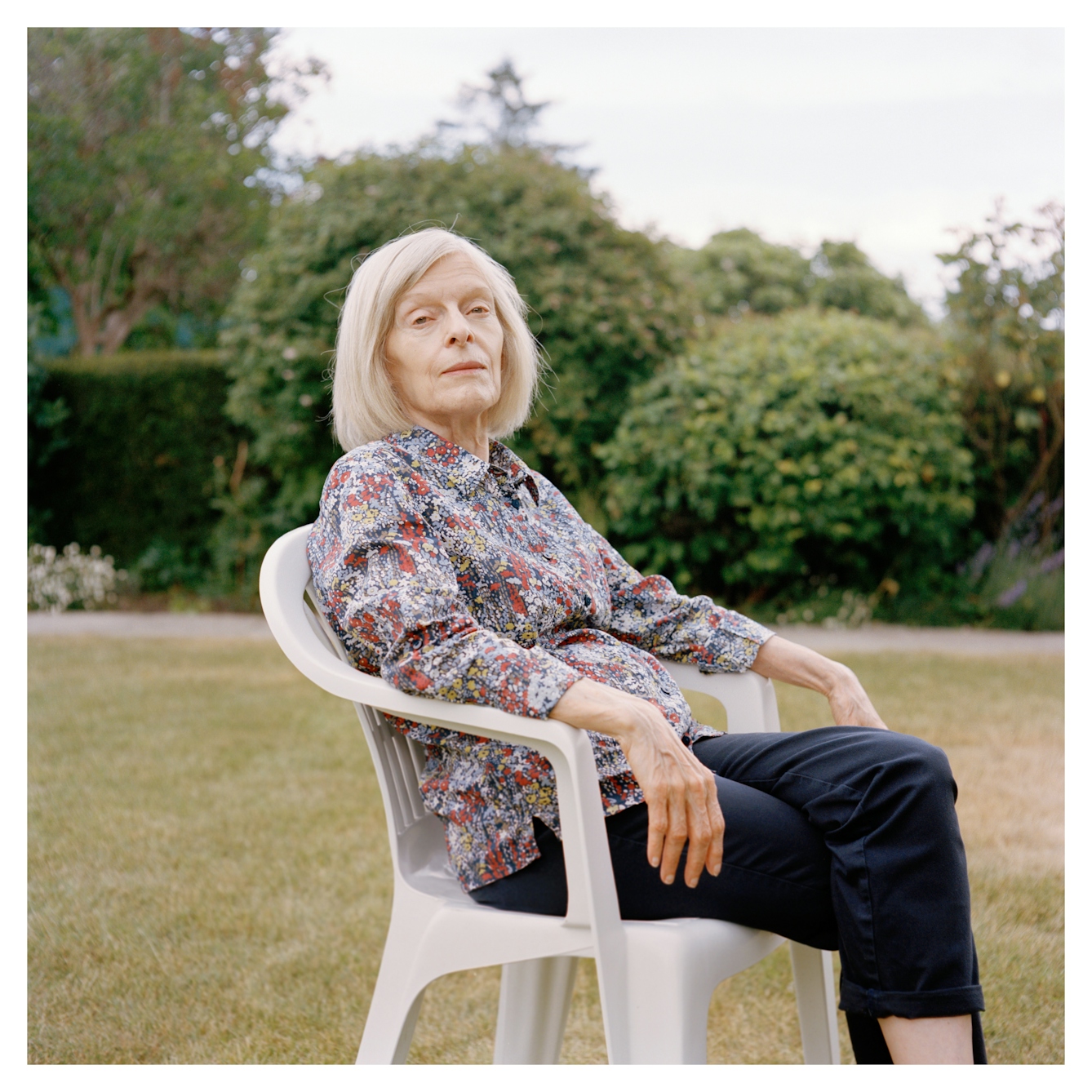 Portrait photograph of Margaret, an older woman with grey shoulder length hair. She is sitting in a white plastic garden chair in a garden.
Her body is sat sideways, with her head turned to face towards the camera. Her arms are resting on the chair arms.
She is wearing a floral patterned shirt and black trousers. She is looking directly at the camera, with a neutral expression.