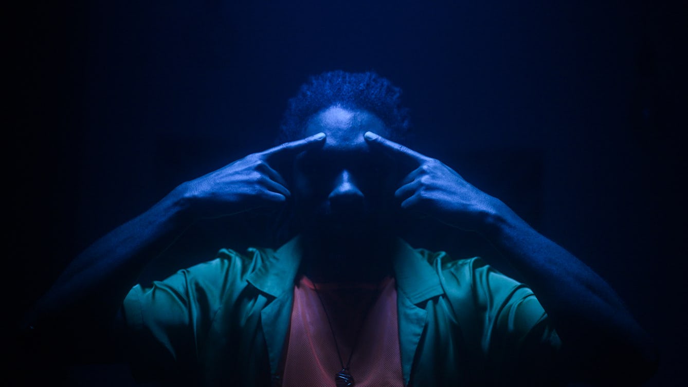 Film still of a man in a dark space, lit with a single blue light from directly above his head, casting much of his face into shadow. He is only visible from the chest up. He is wearing a green shirt, a light red t-shirt and a chain necklace. Both his arms are raise with both his index fingers pointing to his forehead, just above his eyebrows.