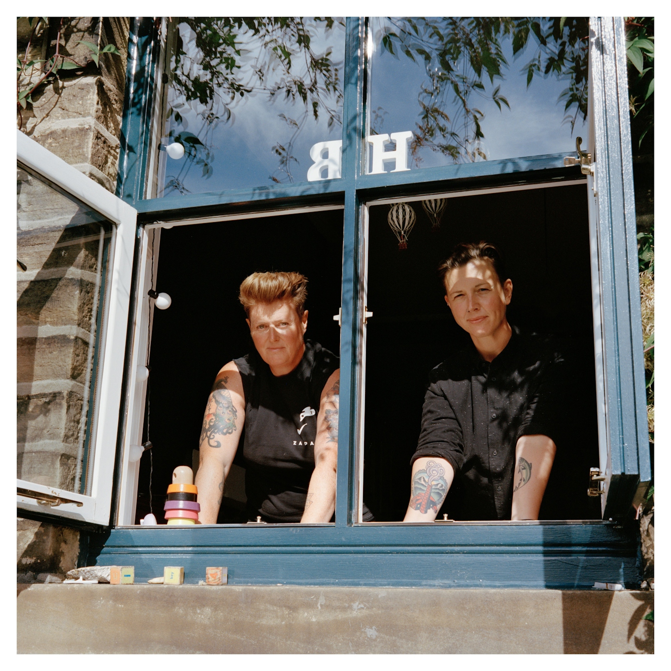 Portrait photograph of Belinda and Heidi standing in an open window, looking outside towards the camera. The woman on the left has short brown hair and is wearing a black sleeveless top, she has several tattoos on her arms. The woman on the left has short dark hair and is wearing a black shirt - she has several tattoos on her arms.
They are both looking directly at the camera. There is a stack of colourful wooden rings on the windowsill and above their heads, a wooden initial H and B.