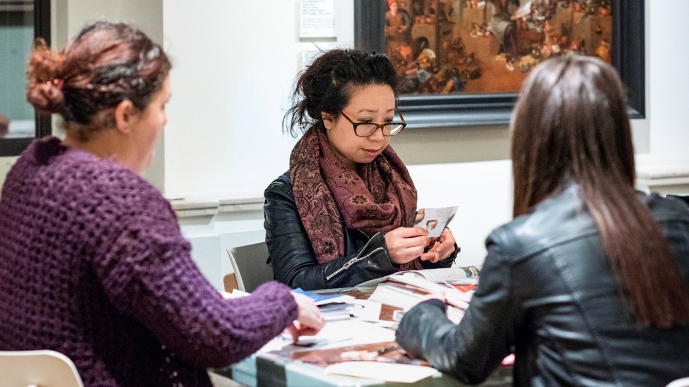 Photograph of three people sitting at a table looking at materials for a workshop in Wellcome Collection. One person is cutting out a page from a magazine.