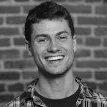 Head and shoulders photograph of a man with shirt hair and even teeth, smiling, wearing a chequered shirt over a black t-shirt. 