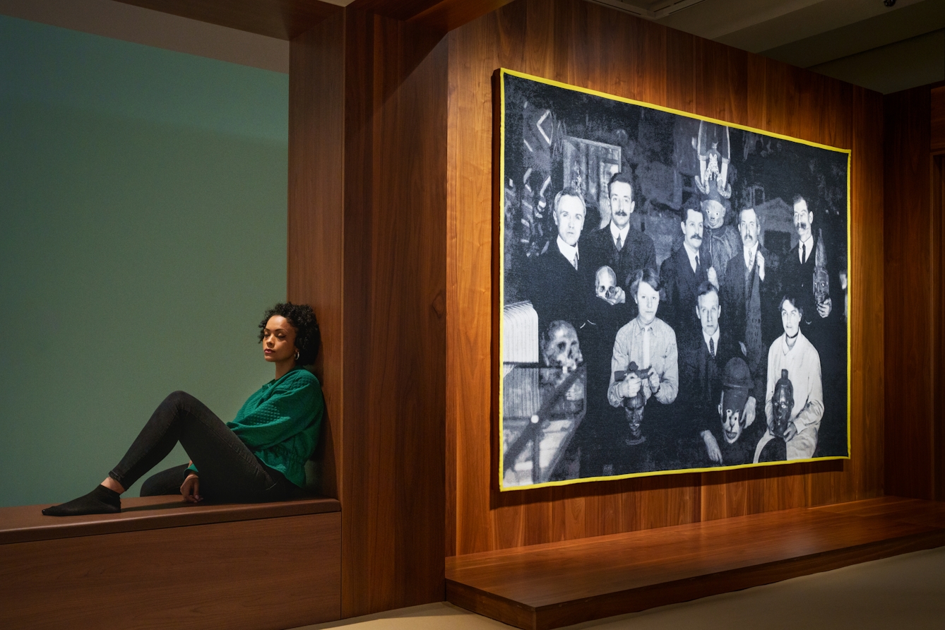 Photograph of a gallery exhibition space showing large tapestry hung on a wooden wall. The tapestry is made up of a black and white, archive photograph, from the early 20th century showing a group of white men and women gathered in a group holding museum artefacts in their hands or on their laps. They are surrounded by other museum exhibits and display cases. The tapestry is framed with a bright yellow border. Surrounding the tapestry is a wider wooden structure containing seating and alcoves. To the right of the image a gallery visitor reclines in one of the alcoves with her back to the tapestry, eyes closed in contemplation. 