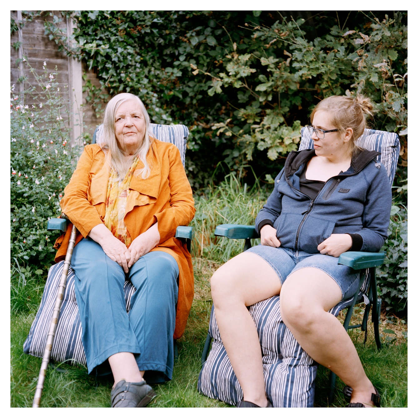 Portrait photograph of two women, Alison and Lex, sitting close to one another on plastic garden chairs in an outdoor setting. Alison is a woman with long grey hair, wearing blue trousers, a patterned top and an orange jacket. She has a silver walking stick which is leaning against the edge of her chair. She is looking at the camera. Lexy is sat beside her. She has blonde hair tied in a bun. She is wearing a blue zipped jumper, denim shorts and a black t-shirt. She is looking at Alison and smiling slightly, the corner of her lips are turned upwards. Behind them are are some trees and small plants, along a brick wall.