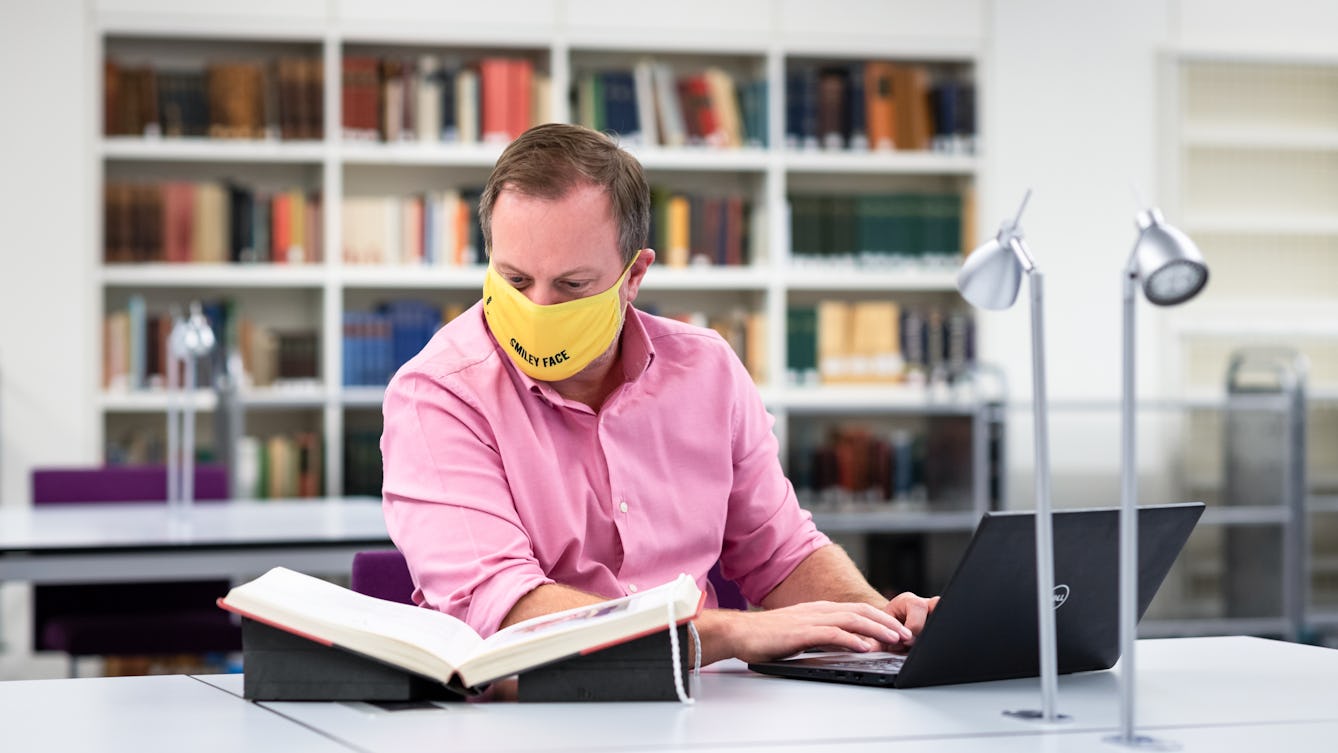 Photograph of a man wearing a pink shirt and a yellow face covering sitting at a table in a library. His hands are wresting on the keyboard of a laptop, whilst he looks at an open book, resting in a book cradle. Behind him in the distance a book shelf and other tables can been seen.