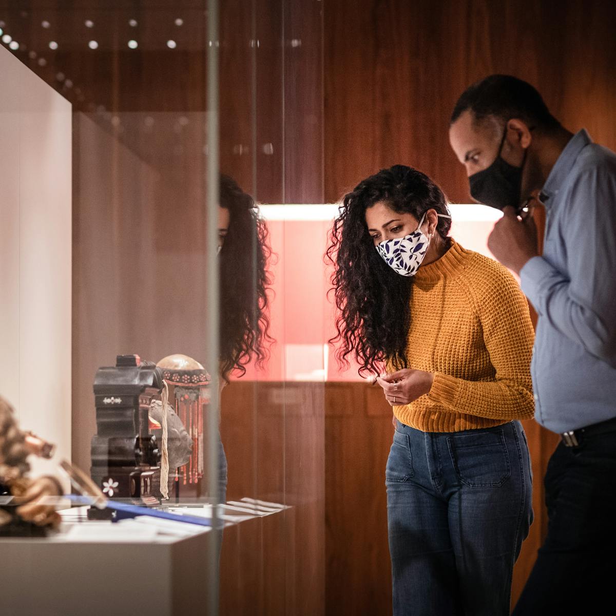 Photograph of a man and a woman wearing face coverings exploring a museum gallery. The woman is wearing a yellow knitted jumper and a pair of blue jeans. The man is wearing a blue shirt and dark trousers. It is a dark gallery with spot lit exhibits and wooden panelled walls. The couple are standing side by side to the right of the frame in mid discussion about objects in a display case. To the left of the frame is the large glass display case they are looking at, containing several objects, from vases to models of the human head to small cabinets.