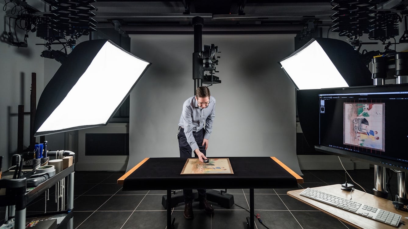 Photograph of a windowless photographic studio. In the centre is a table covered in a black cloth. Standing over the table is a man holding a light meter above an artwork which is resting on the table. Behind him is a large camera stand. To either side are large softboxes mounted on studio flash heads. In the foreground to the left is a trolley containing studio accessories. In the foreground to right is a computer display and keyboard.