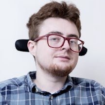Photograph of a white person with short brown hair and a short beard. They are wearing glasses and a checked blue shirt.