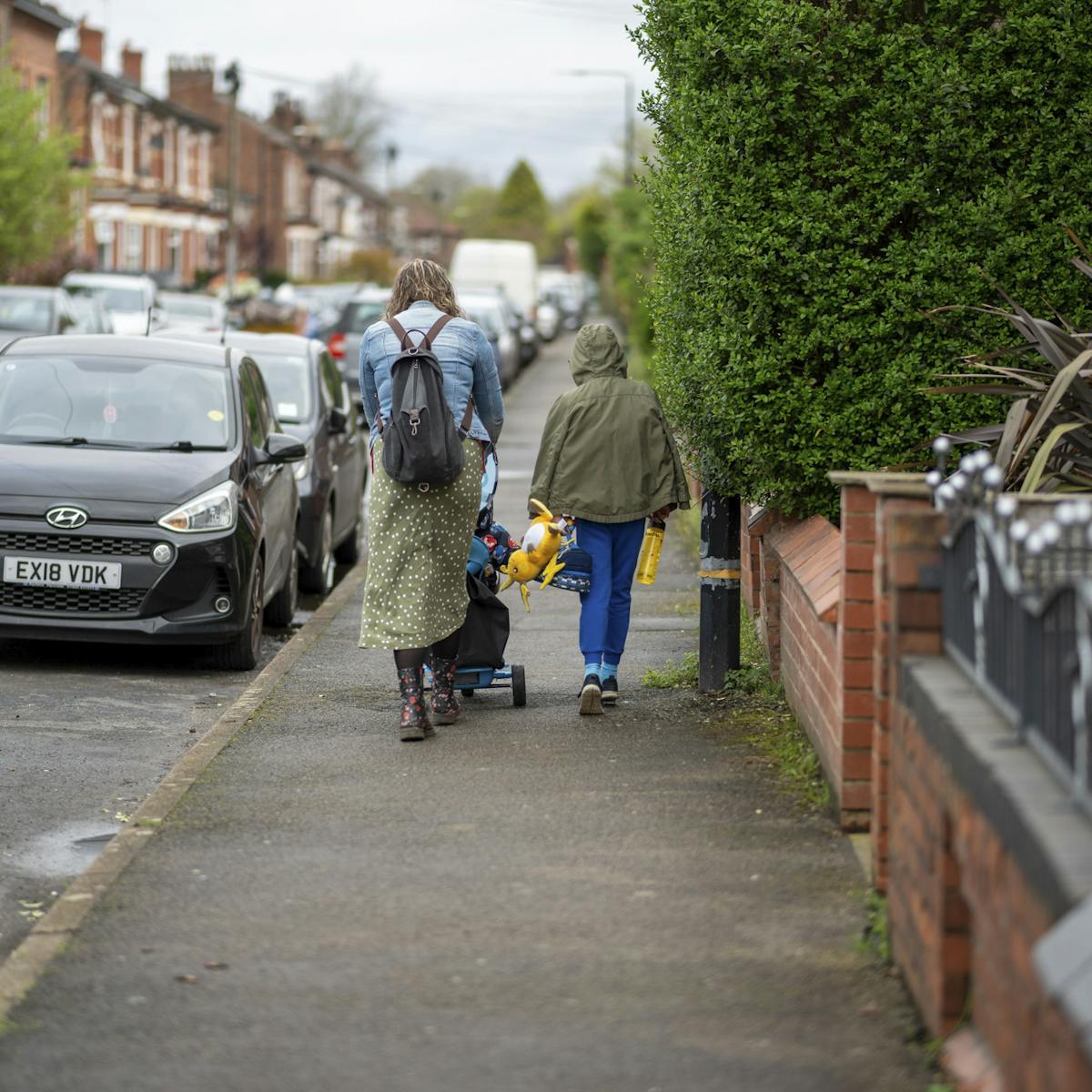 A photography of a mother and her two children walking on the pavement next to the road. They are walking away from us, the older child walks alongside the mother, who is pushing a pushchair with the younger child.