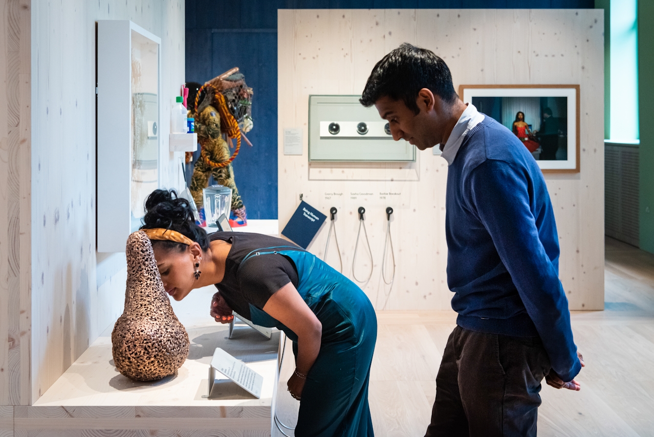 The photo shows a visitor bending down to smell a a bronze gourd shaped sculpture, the texture of which is like a porous rock. The sculpture is sitting upright on a wooden panelled top and surround.