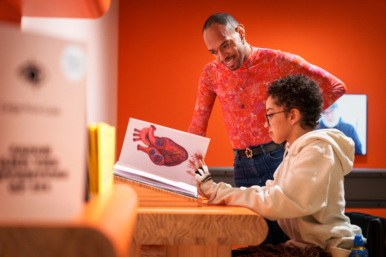 Photograph of two gallery visitors leafing through an artist's sketchbook resting on a table. On the half turned page, a sketch of a large colourful anatomical heart can be seen.