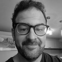 Black and white photograph of the head of a young white man with short dark hair and large black-framed glasses. He is smiling and looking into the camera.