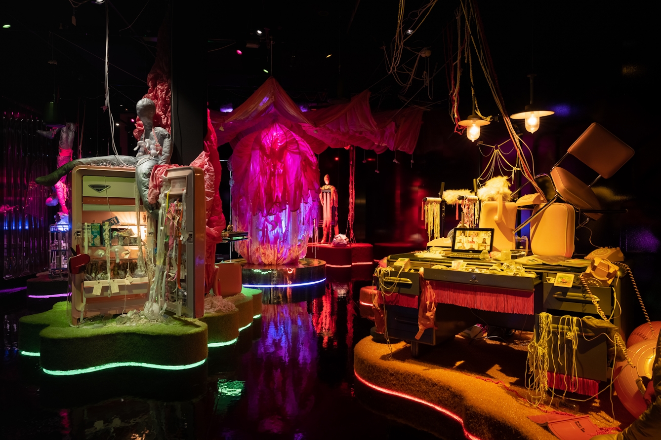 Photograph of an art installation in gallery space. It's a dark space with black glossy floors and walls. Floating on island-like structures within the space are vibrant coloured lights of greens, purples, oranges and reds. On these structures are busy vignettes, a fridge with it's door open and reams of tape spilling out, a large parachute suspended from the ceiling and a large desk covered in a laptop, telephones and wires.