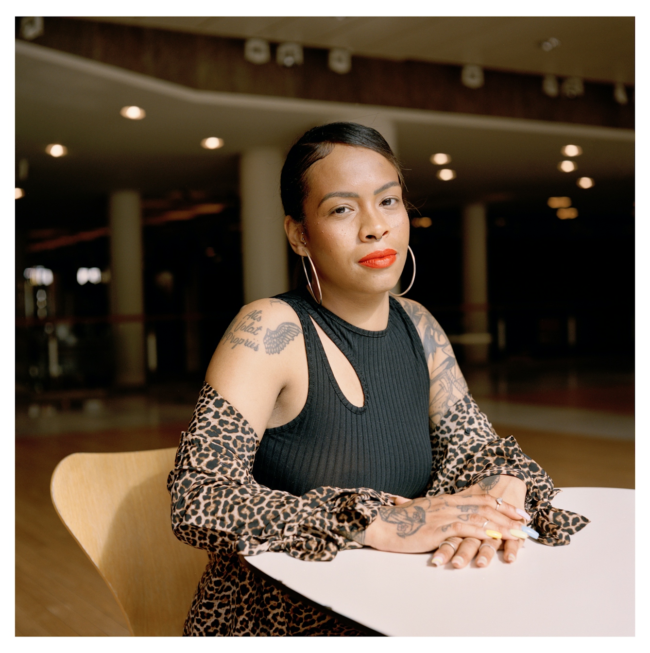 Portrait photograph of Lauren, a young Black woman, sitting on wooden chair. She has several tattoos on her arms and is wearing a black sleeveless top and a leopard print skirt. She has large hoop earrings and is wearing an orange red lipstick.
She is looking straight to camera with a quiet smile. Behind her, there is an exhibition space which is empty apart from several wooden chairs.