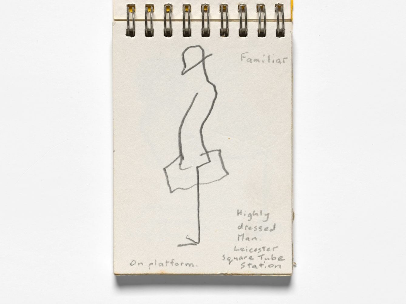 Photograph of an open sketchbook on a white background. A simple line sketch is visible, showing a man in a hat, carrying a case, stood on a station platform. Although only made up of a few lines, the sketch conveys a sense of waiting and contemplative thought. The sketch is hand annotated with the words, 'Familiar, Highly dressed man. Leicester Square Tube Station. On platform."