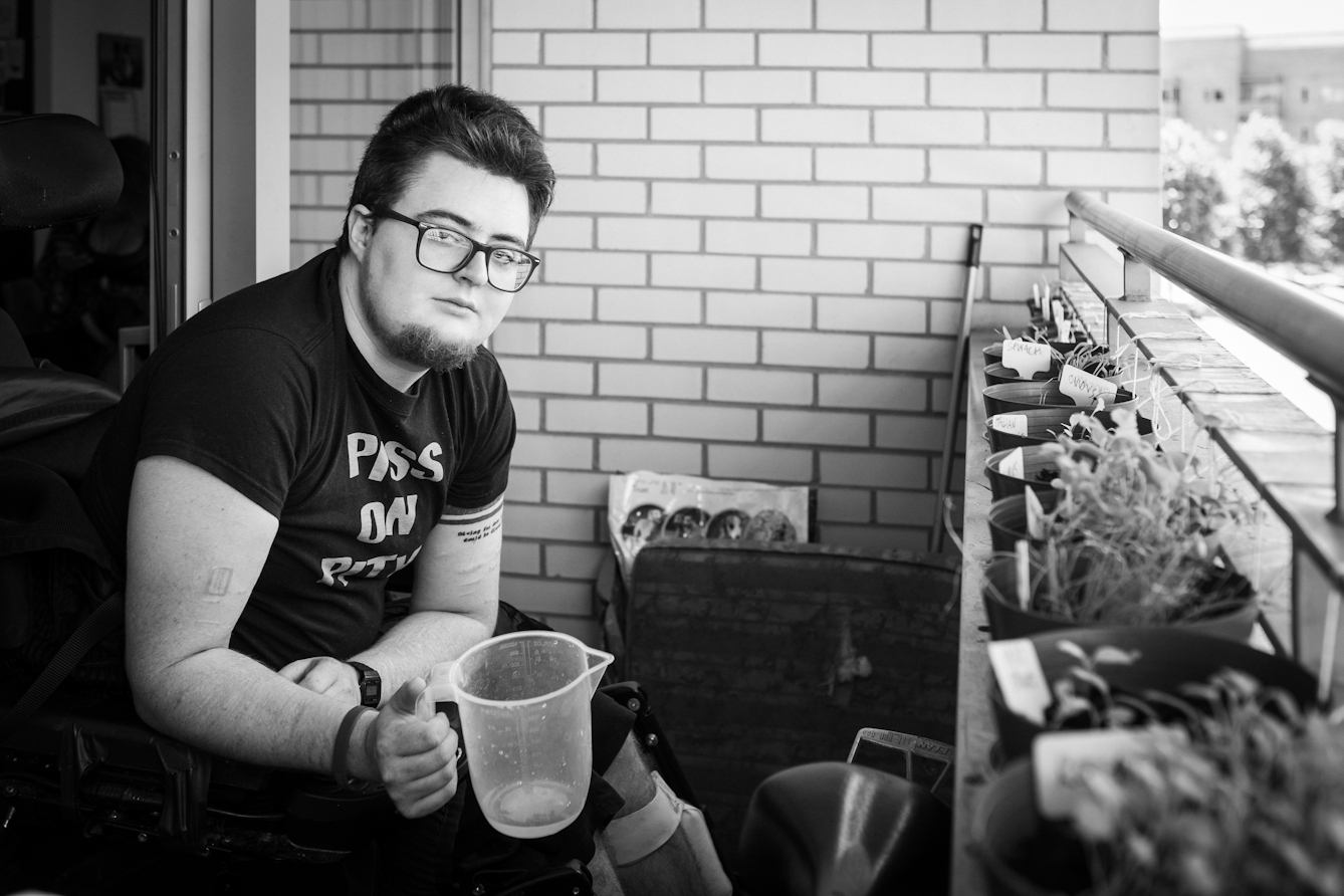 Black and white photographic portrait of Jamie Hale on his balcony at home watering plants.